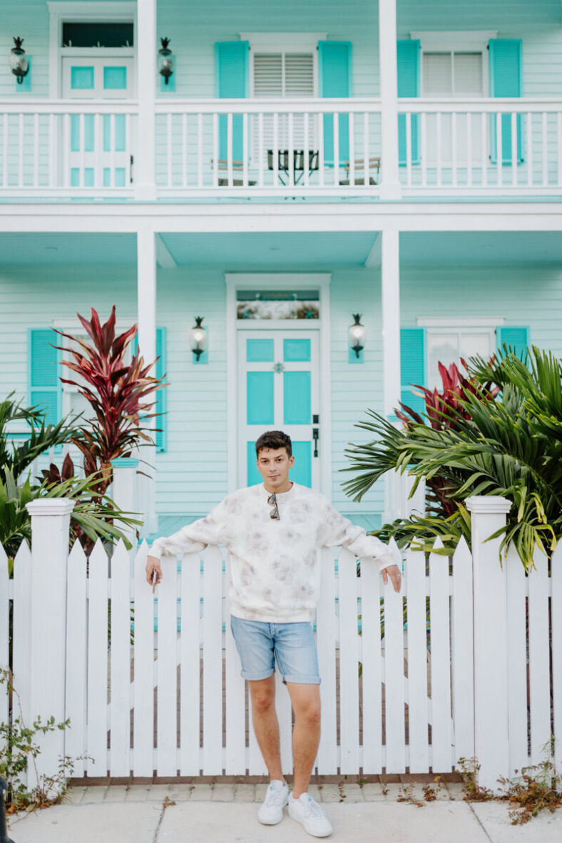 A man in a sweater stands in front of an aqua colored house with white trim.
