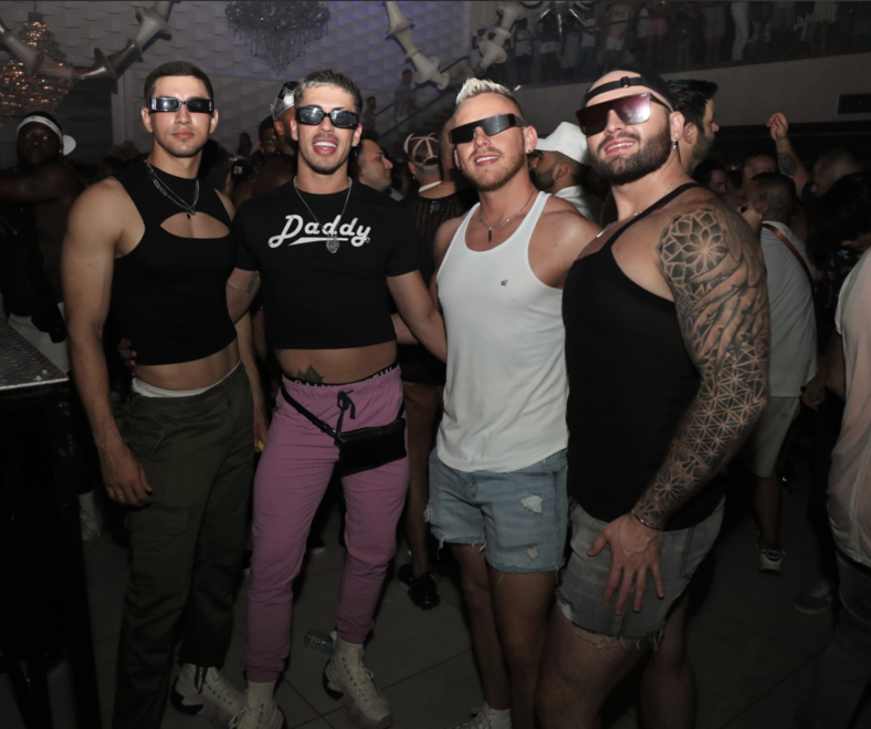 Four gay men in sunglasses and warm-weather apparel stand and pose for a picture together at teh White Party in Fort Lauderdale