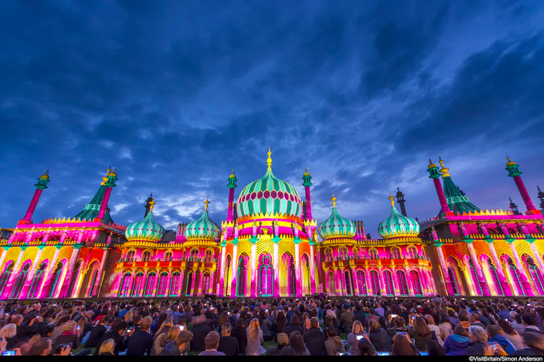 Crowd of people in front of the Royal Pavilion at dusk, lit up in a range of vivid colours, pink, green, yellow and purple, Brighton, East Sussex, England.