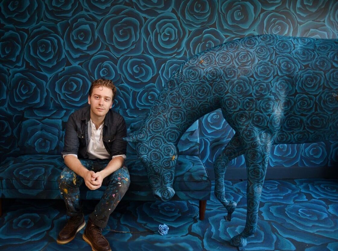 Wiley Caudill sits in front a patterned wall next to a horse statue. The wall, floor and horse are all overed in painted blue roses.