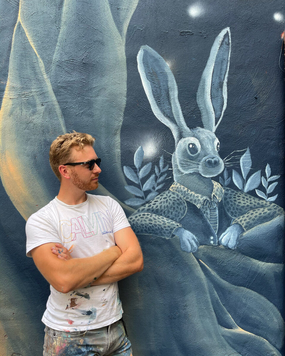 Caudill, wearing paint stained clothing, shows off another of his murals that features a rabbit wearing a shirt and vest.