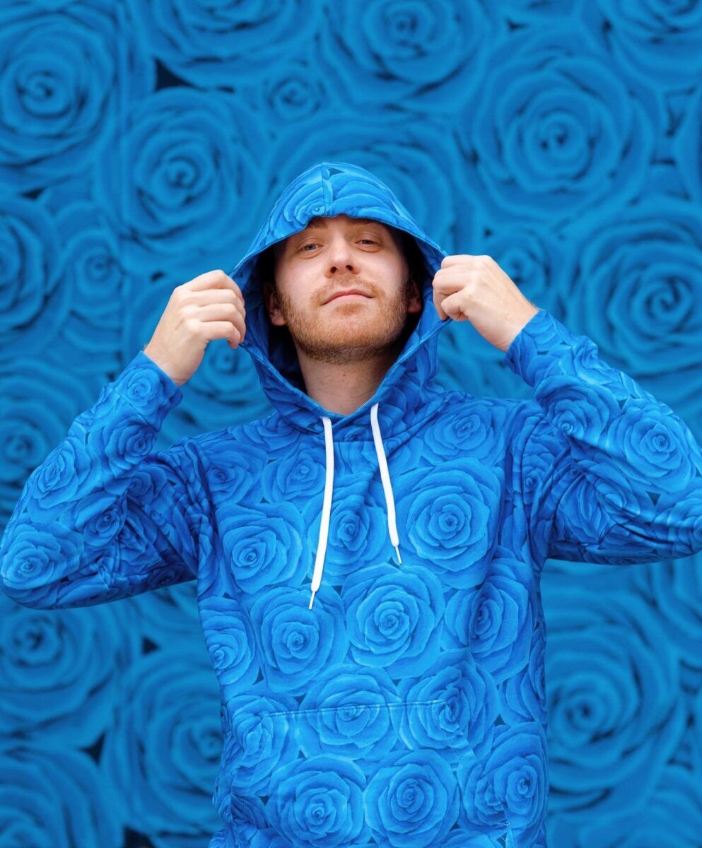Caudill poses in front of a wall featuring his trademark blue roses. He is wearing a hoodie with the same rose motif.
