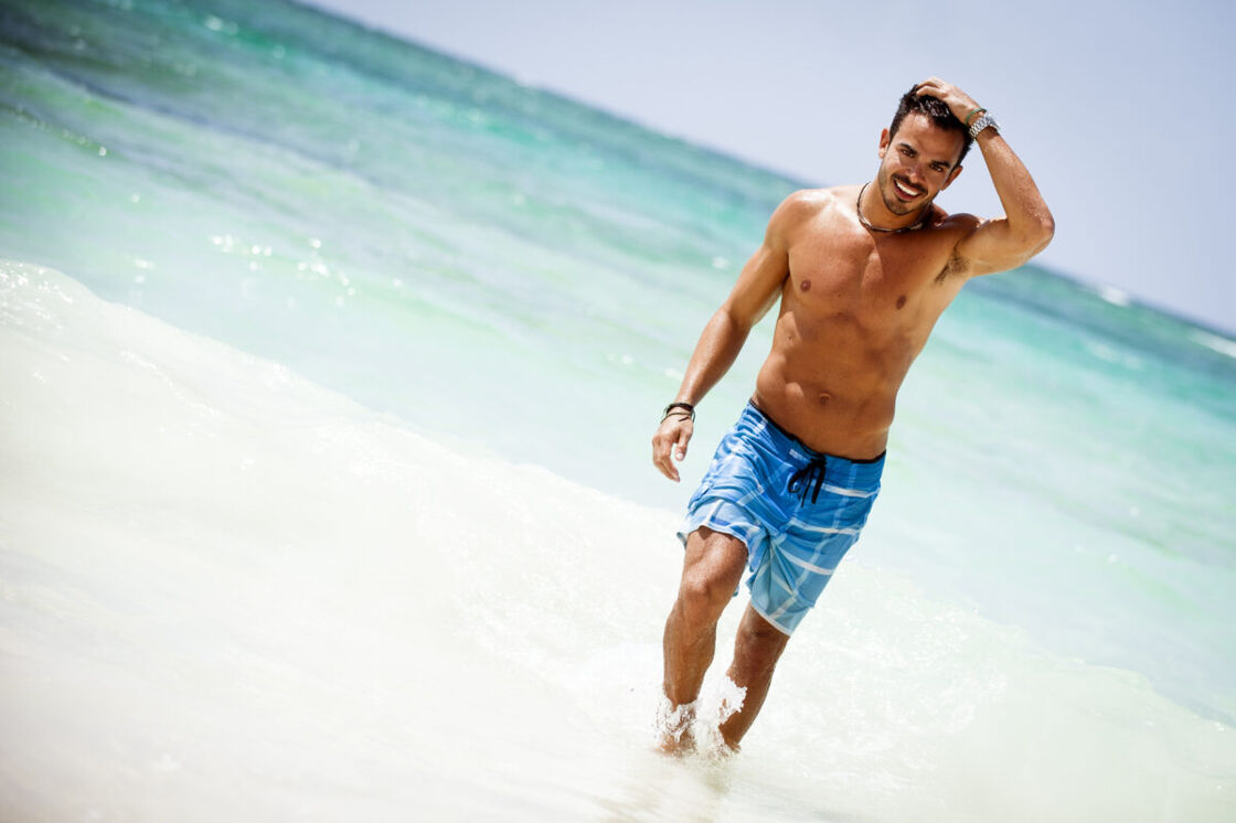 An attractive young gay man wearing swim trunks on the beach by the ocean of an LGBTQ+-friendly Caribbean island