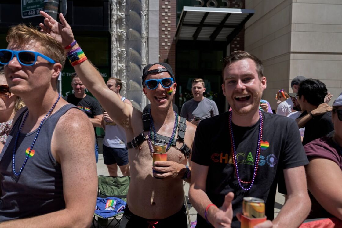 A group of cheerful people celebrating at a pride event, with one man wearing a black "Chicago" t-shirt with a rainbow emblem, and another shirtless with a harness and sunglasses, holding a drink.