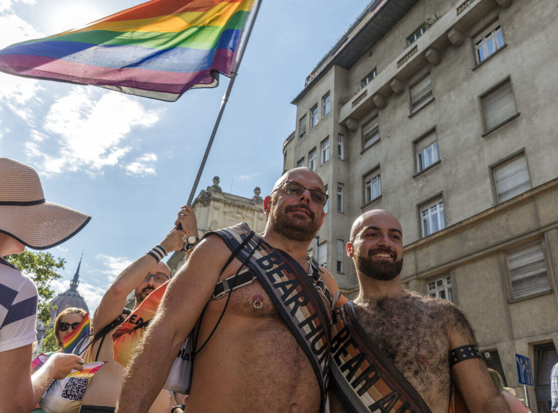 Two gay, European bears wearing their leather Mr. Bear sashes at a Pride event with rainbow flags