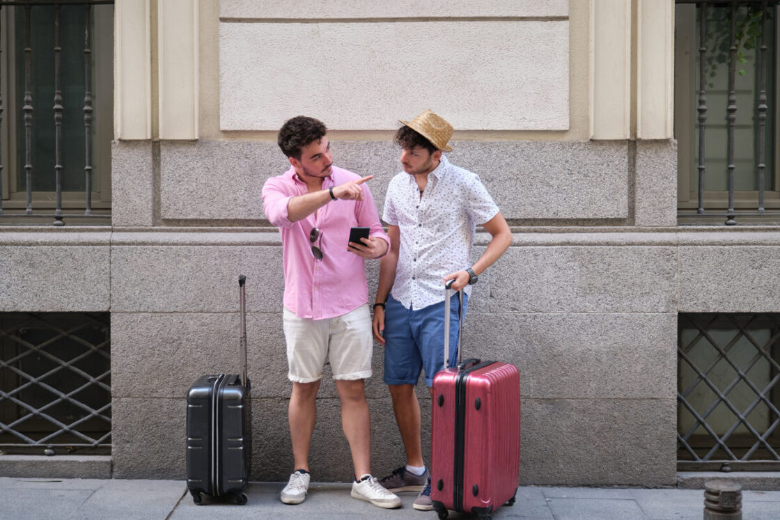 Two men stand together on a city sidewalk, possibly tourists, each with a suitcase. The man on the left, wearing a pale pink button-up shirt and beige shorts, points to something out of frame with his right hand while holding a smartphone in his left. The man on the right, in a white patterned shirt, navy shorts, and a straw hat, looks at the phone screen with a focused expression. 