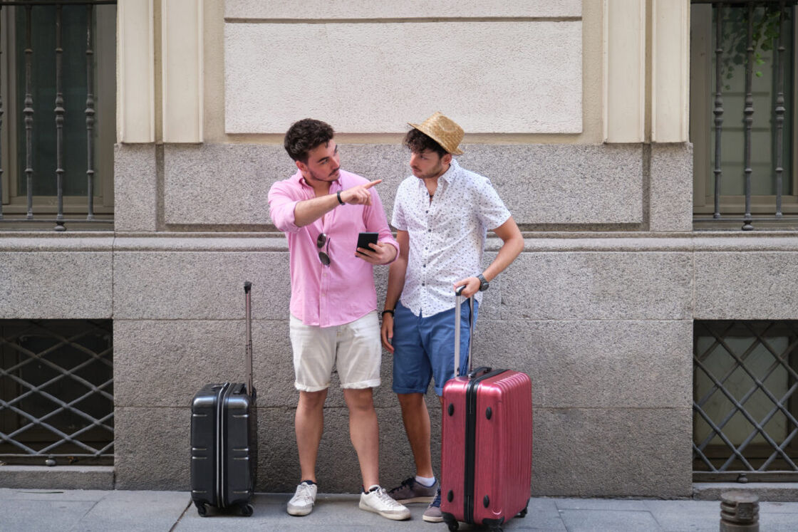 Two men stand together on a city sidewalk, possibly tourists, each with a suitcase. The man on the left, wearing a pale pink button-up shirt and beige shorts, points to something out of frame with his right hand while holding a smartphone in his left. The man on the right, in a white patterned shirt, navy shorts, and a straw hat, looks at the phone screen with a focused expression.