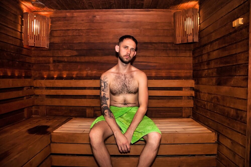 Young man in green towel sitting on bench in bathhouse sauna and relaxing, looking at camera.