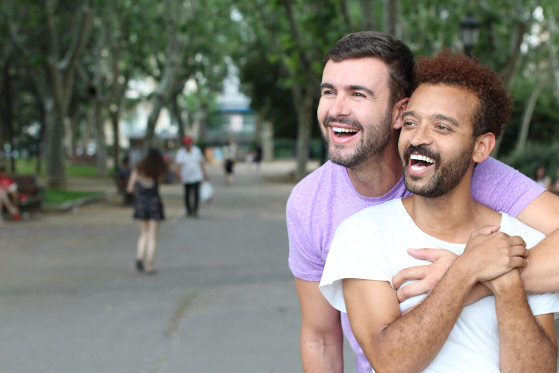 Two queer men are standing close together in what appears to be a joyful embrace in a park setting of a gay destination. The man in the foreground is wearing a white t-shirt and has a bright smile, his teeth showing, and curly hair. The man behind him, wearing a purple t-shirt, has his arms wrapped around the other's chest and is also smiling with a look of happiness.
