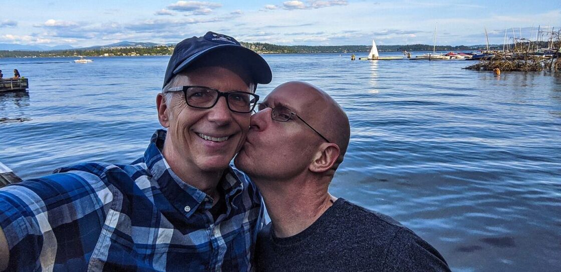Michael and Brent pose for a selfie on the shore of a lake with the water behind them. Brent is kissing Michael on the cheek.