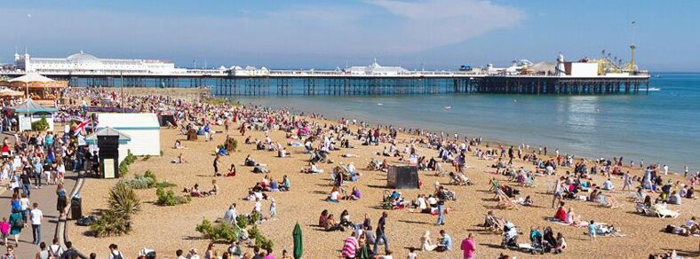 A packed beach on a sunny day with the pier stretching behind. Photo via Visitbrighton facebook