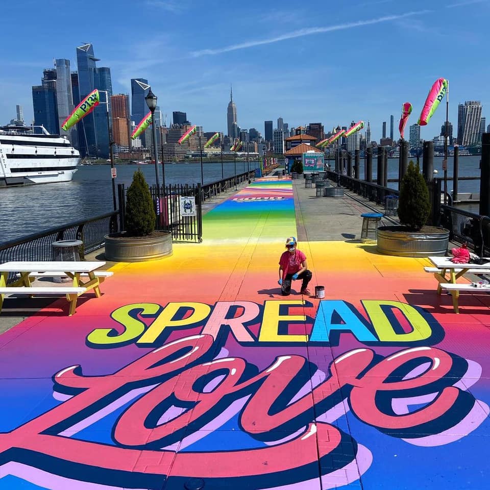 NYC skyline with a rainbow mural with the words "spread love" on the sidewalk. Photo via NYC Pride