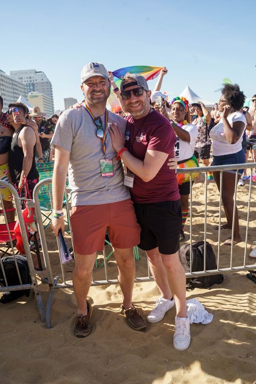 Two smiling men embrace with a crowded beach behind them. Photo via Hampton Roads Pride