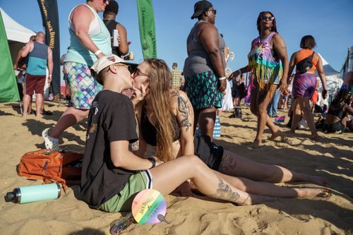 Two female presenting people kiss while sitting in the sand. The beach is crowded behind them. Photo via Hampton Roads Pride