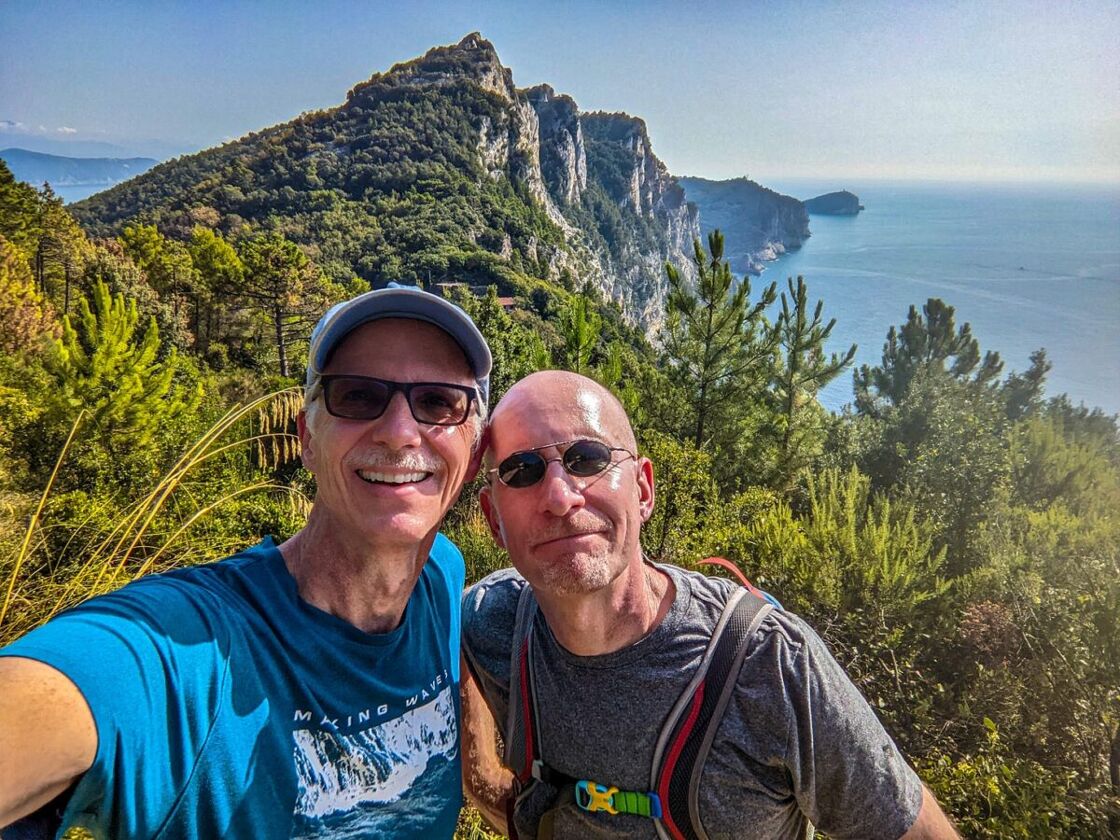 Michael Jensen and Brent Hartinger stand at the top of a hiking trail with the view of the coastline and sea behind them.