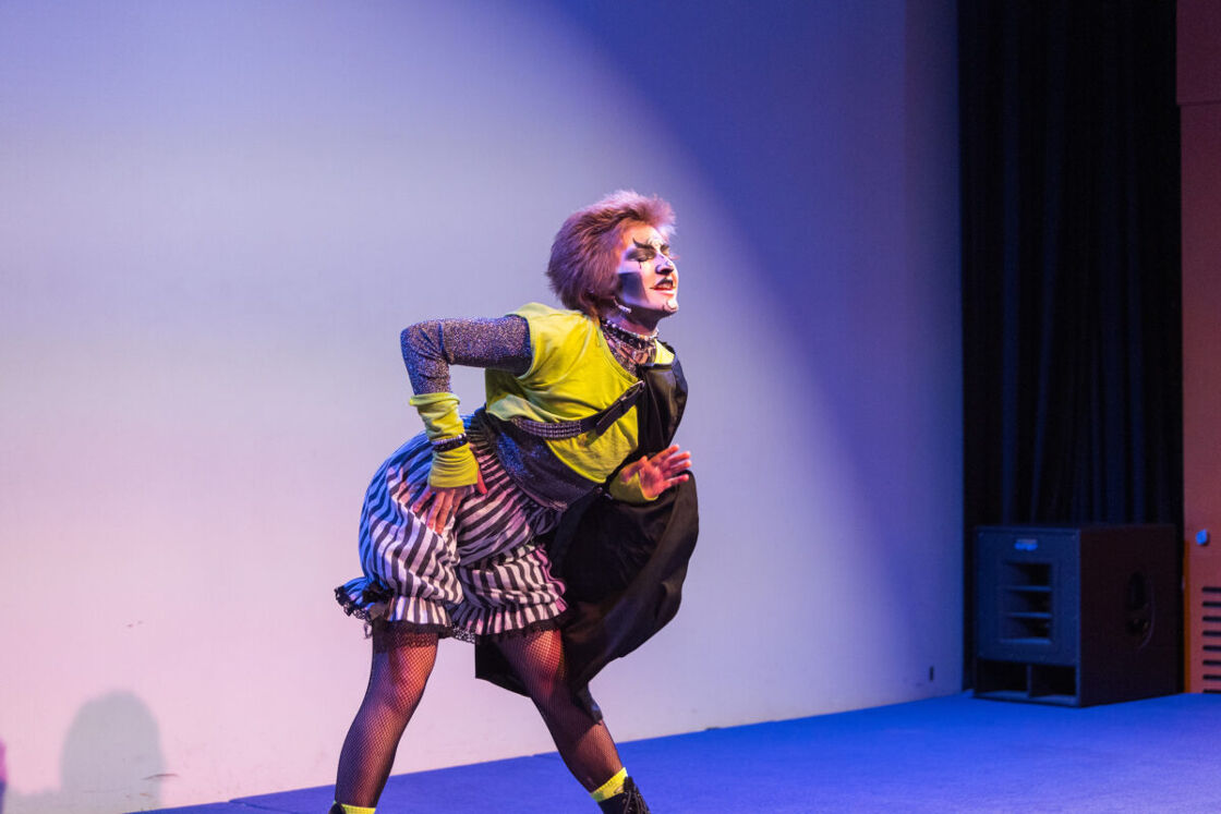 A participant in the School of Drag poses for the cameras. They have on clown makeup, a yellow croptop, and striped shorts.