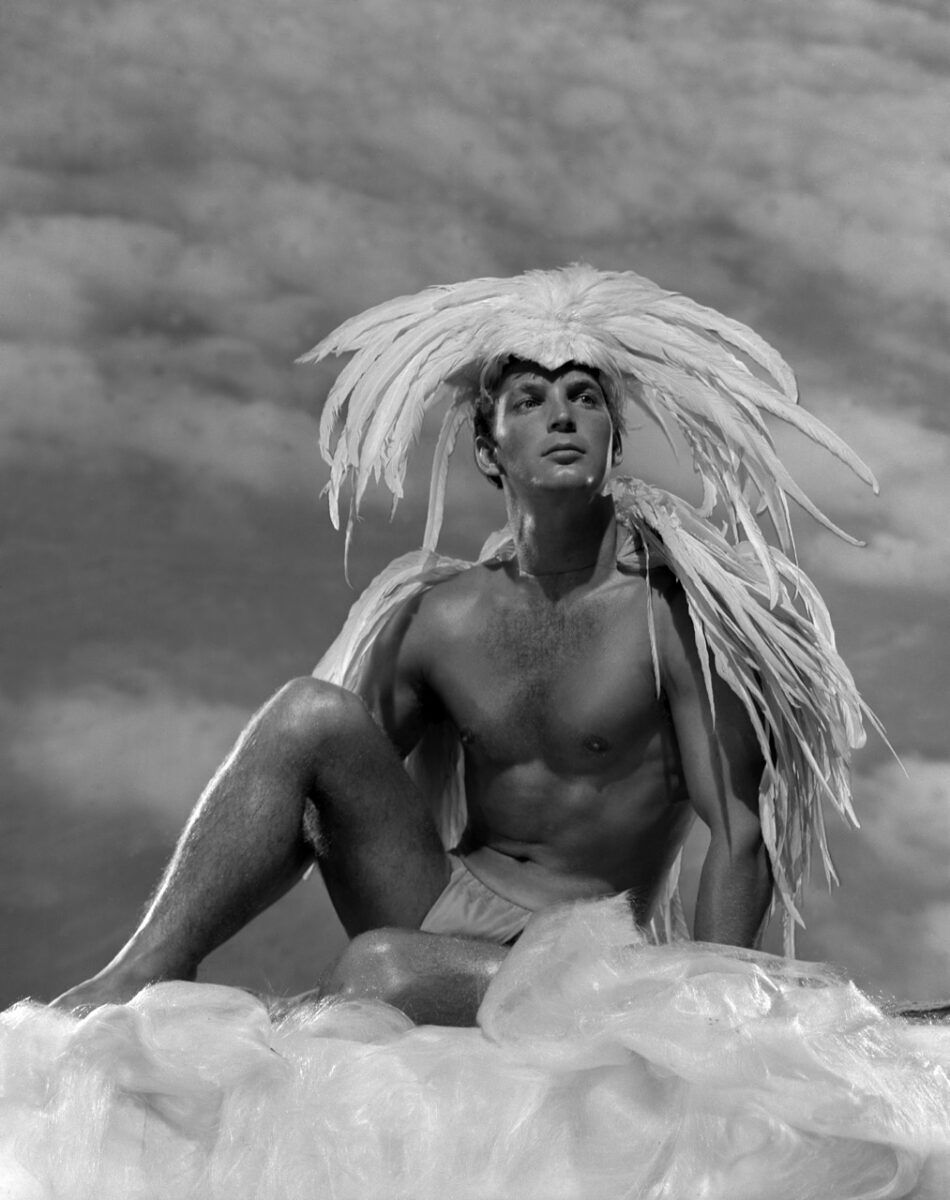 Black and white photo of a young man wearing underwear with feathers on his head