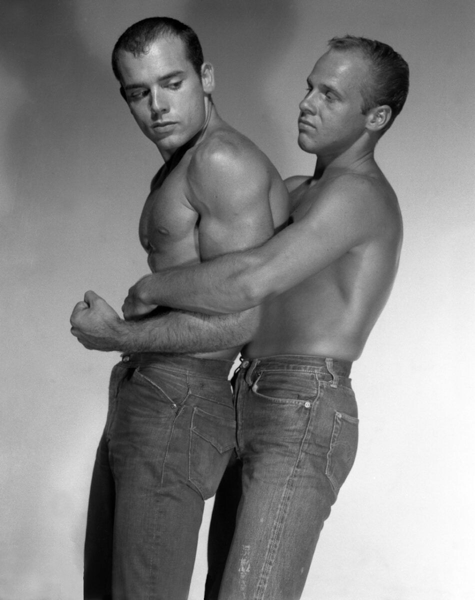 Two shirtless men wearing jeans strike a wrestling pose with one man standing behind the other and pinning his arms to his sides.