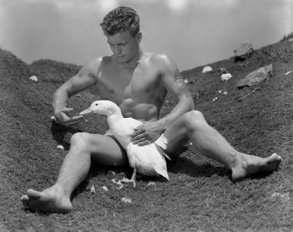 A black and white photo of a young man sitting on the ground with a duck between his legs.