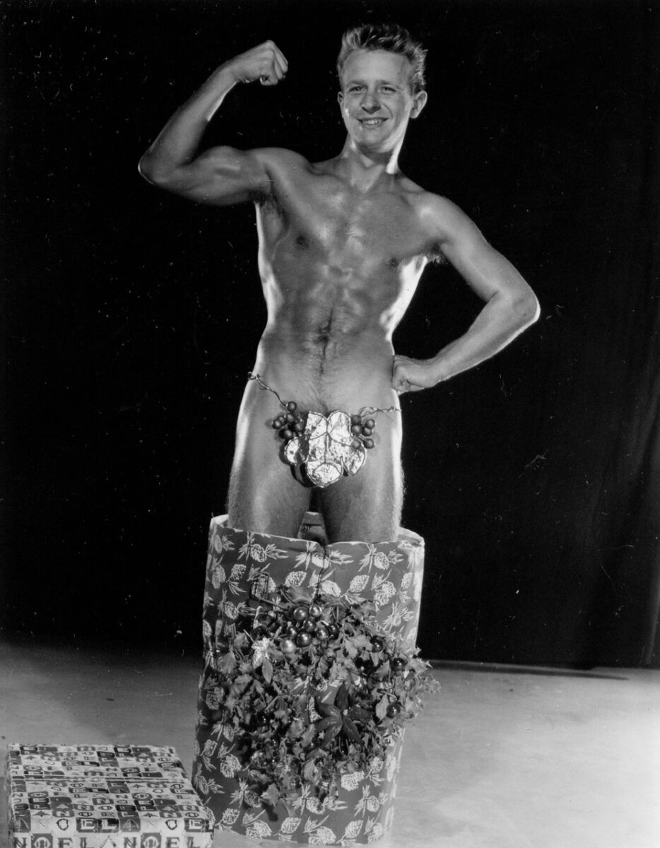 A young man poses as he comes out of a gift package with a bow over his crotch.