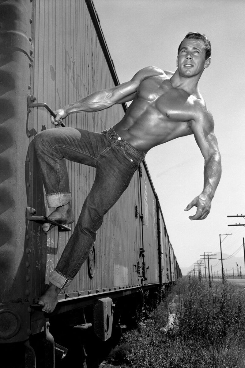 A shirtless young man hangs off the edge of a train