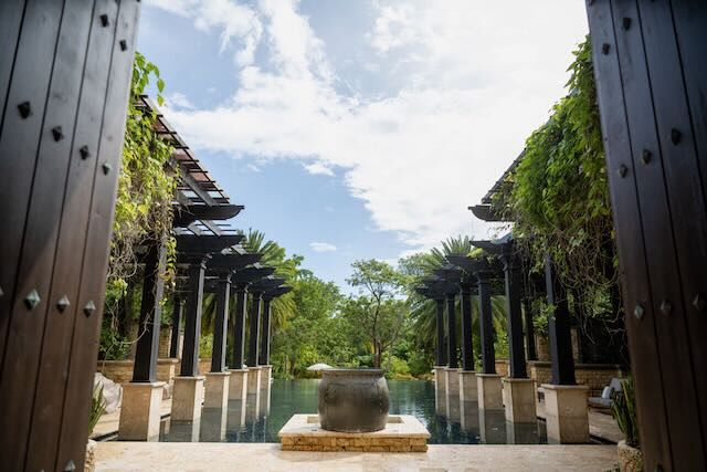 A view from inside a spa of a large quiet pool. A large brown decorative container is in the foreground while pillared alcoves line the sides of the pool