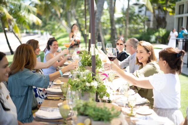 A crowd of people toast each other around a long table with fine linen and china dishes on it. They are outside under palm trees.