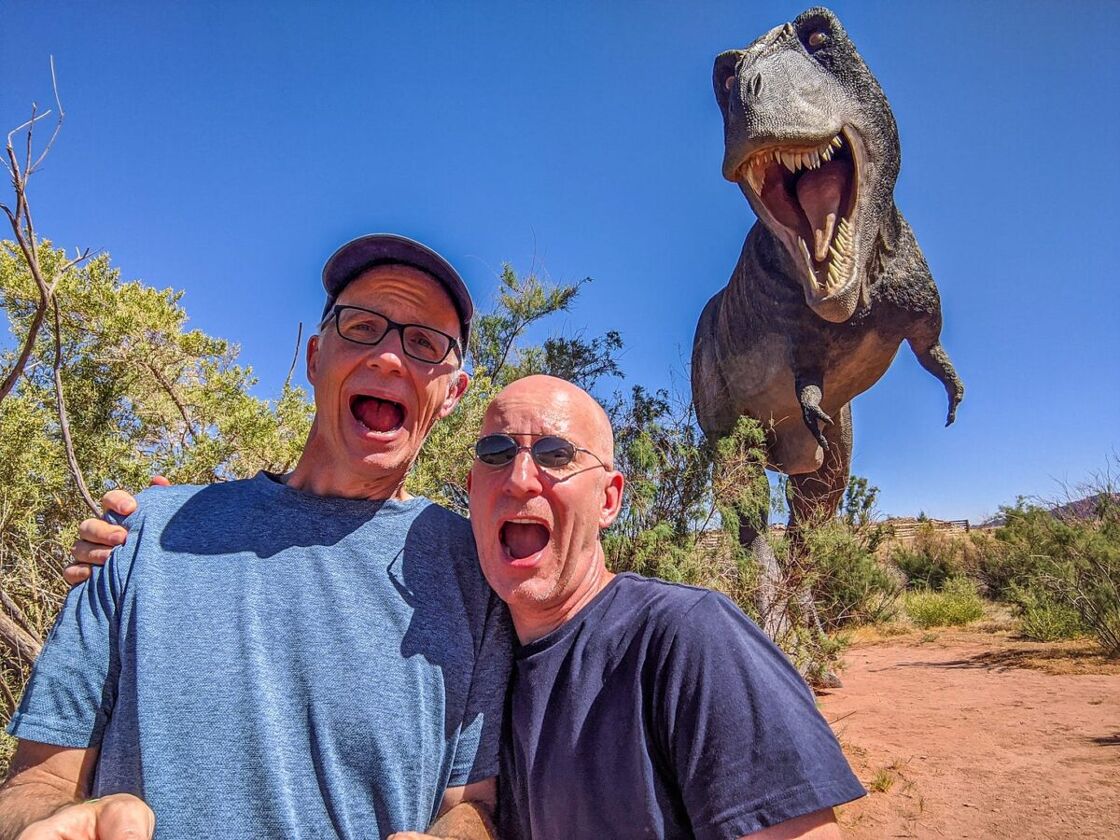 Michael Jensen and Brent Hartinger pose in mock horror in a desert landscape with a statue of a dinosaur behind them.