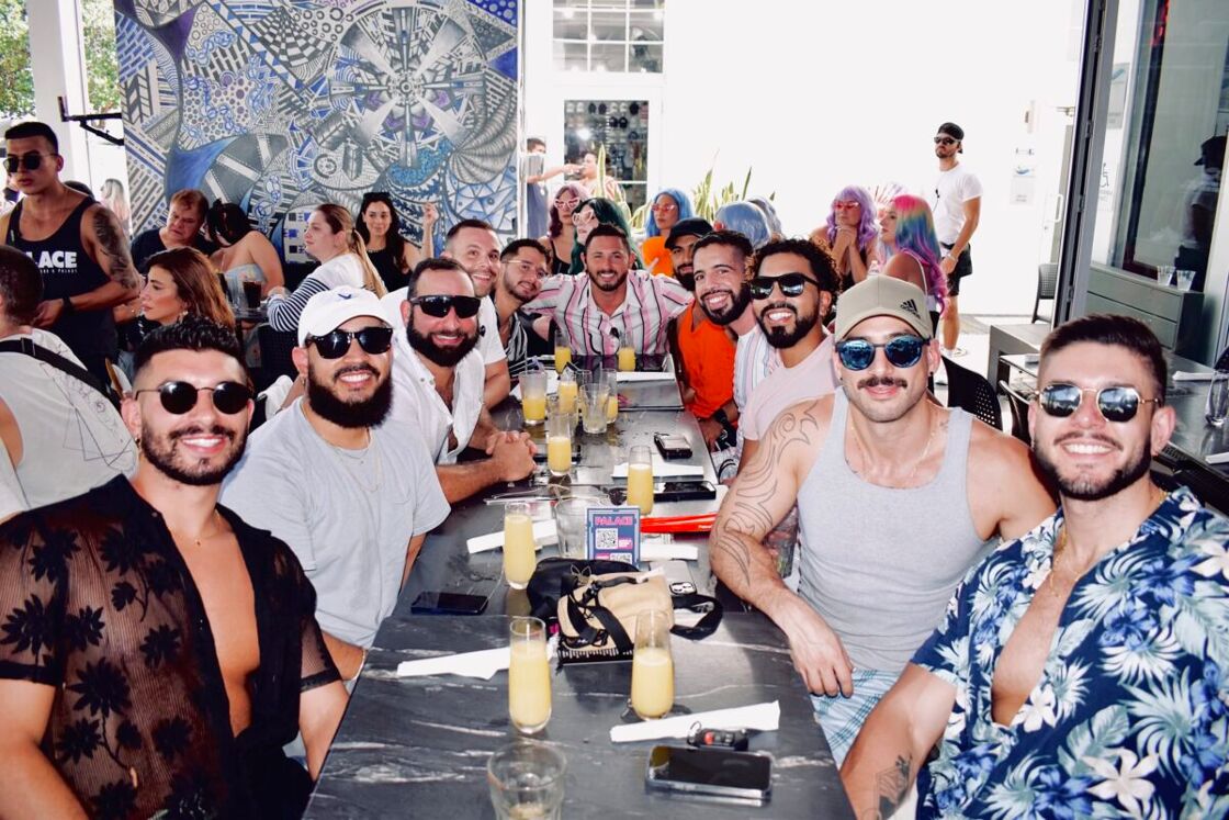 A group of 11 men sit around a table at Palace Bar. Other patrons are in the background.