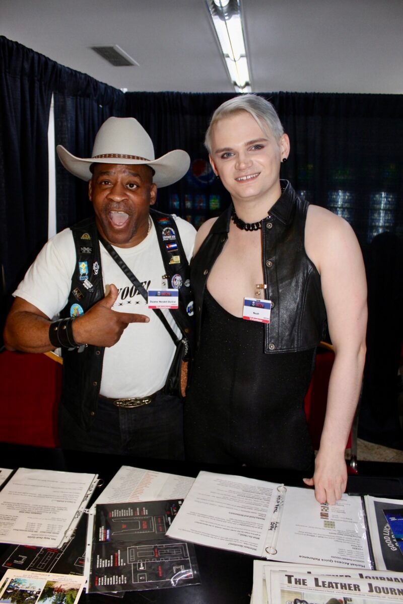 A man and a woman pose for the camera behind a booth at the International Mister Leather conference.