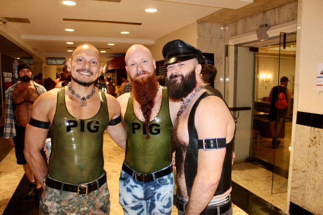 Three men in tight tank tops pose for the camera in a hotel hallway. The shirts say "PIG"