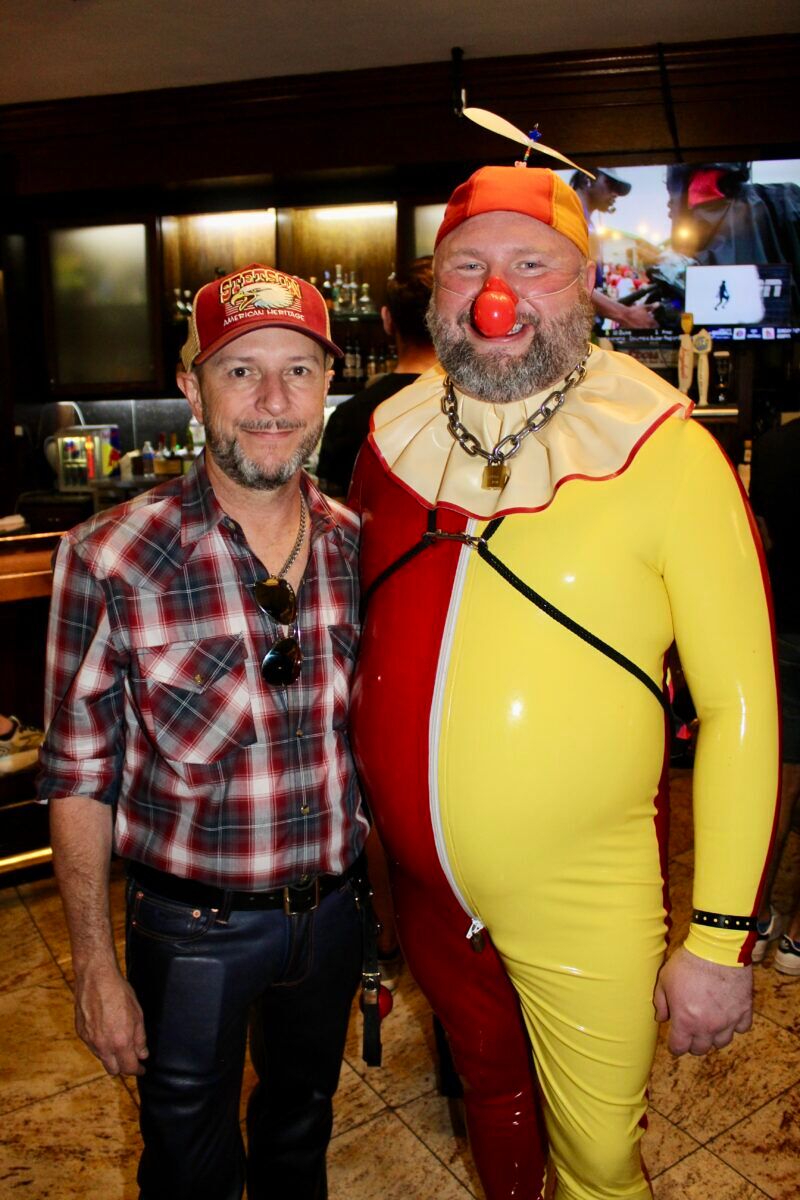 A man wearing a flannel shirts and ballcap poses next to a man dressed in a leather clown outfit.