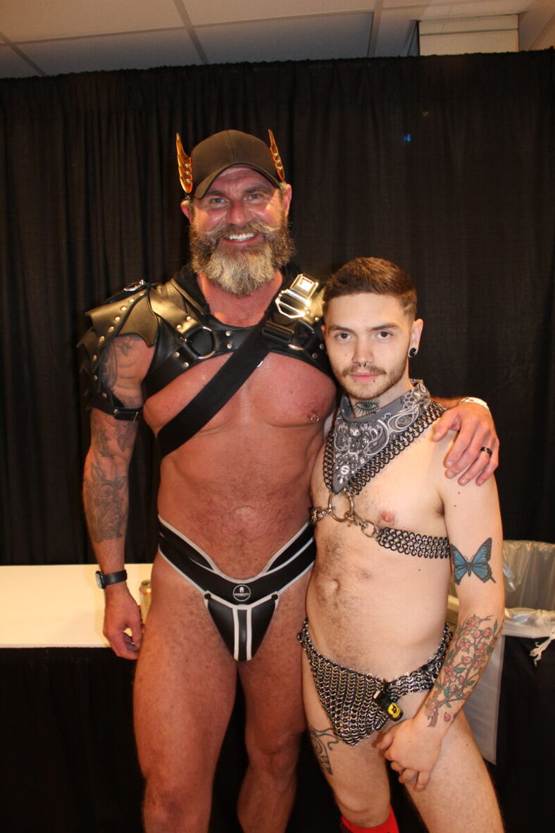 A daddy and his son pose for the camera. Both are shitless and wearing harnesses and jockstraps.