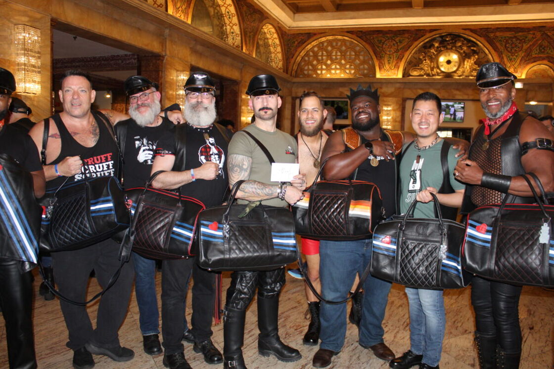 A multiracial group of 8 men pose for the camera with bags emblazoned with a leather flag.