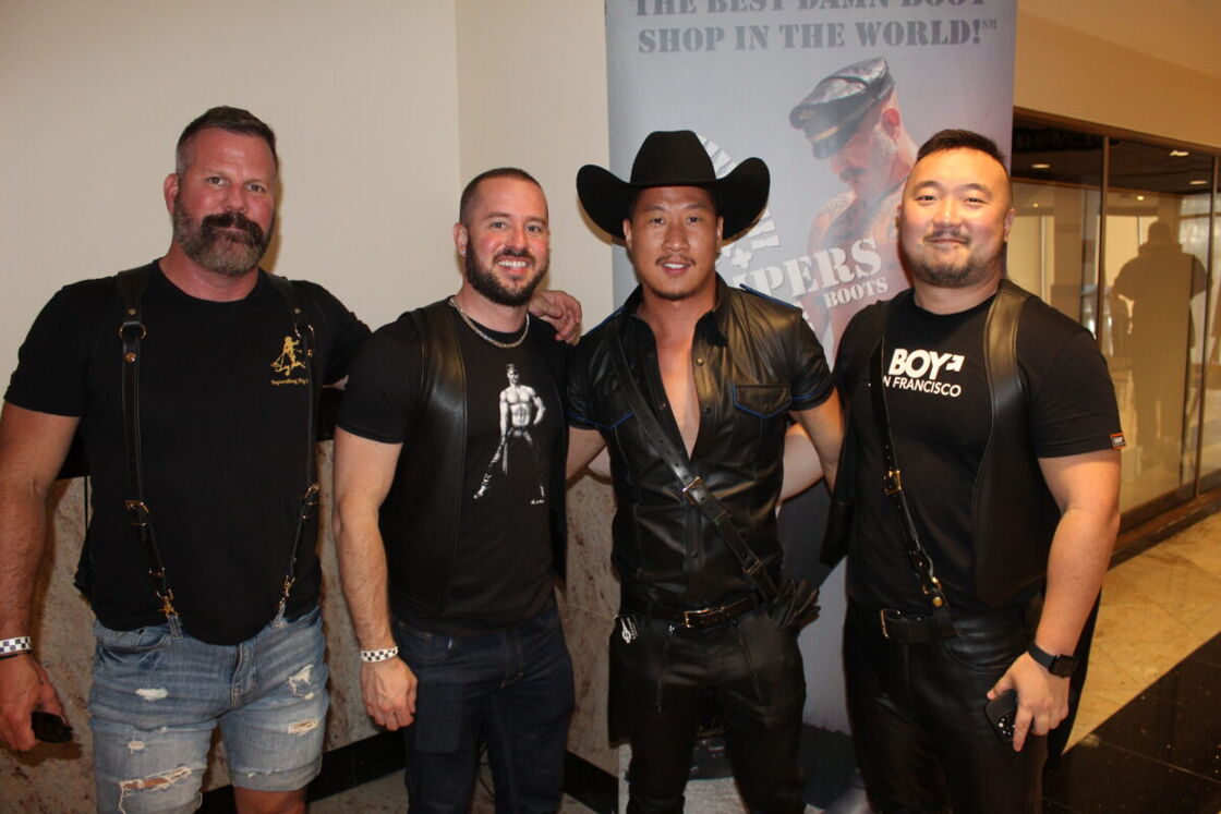Four men wearing all black pose in front of a banner for a boot company.