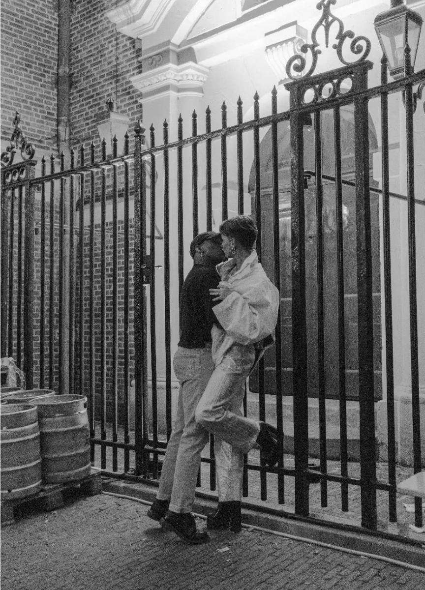 A black and white photograph of an interracial queer couple kiss at the gates by Homomonument.