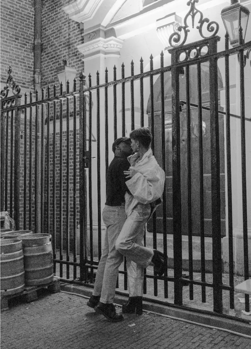 A black and white photograph of an interracial queer couple kiss at the gates by Homomonument. 