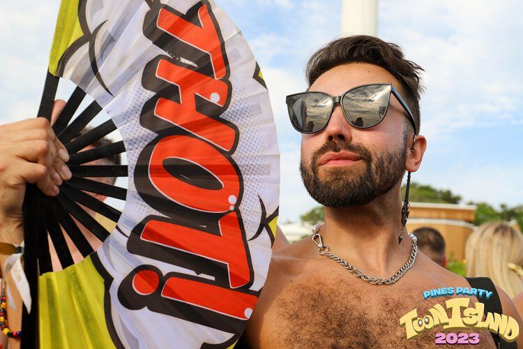 A fabulous bearded man holds a big fan that says THOT! while wearing big reflective sunglasses. Photo via Pines Party