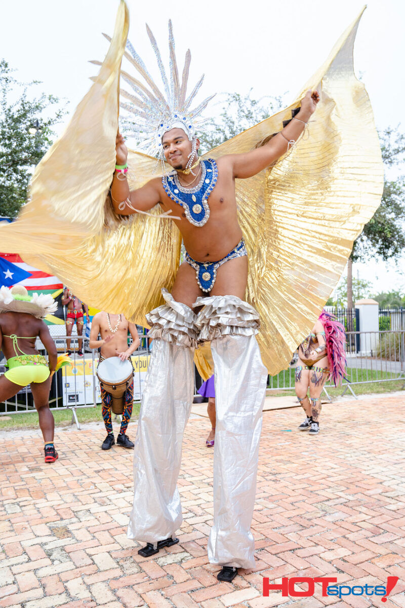 Man on stilts wearing bedazzled jockstrap, headdress and collar is adorned with bright golden wings. Photo via PrideFete
