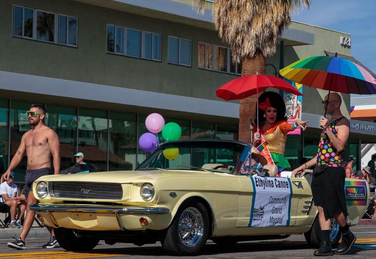 A drag queen sits in a vintage car pointing to the crowd. Photo via https://www.instagram.com/greatautossocal/