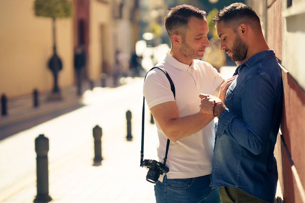 Two men kiss on a deserted street. They are tourists and one has a camera strapped over his shoulder.