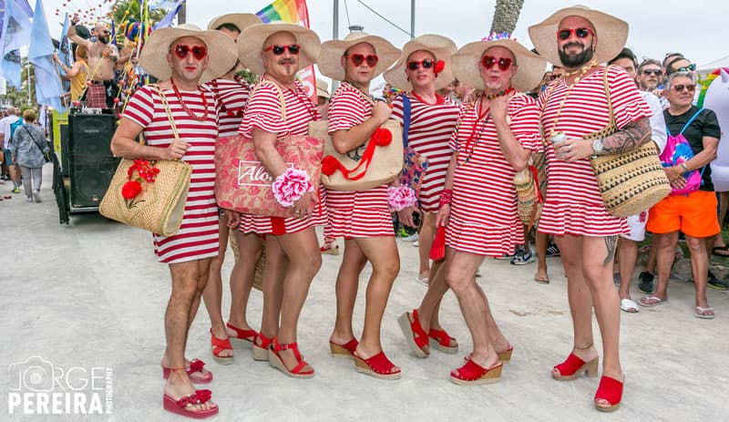 Lovely 'ladies' wearing matching red striped dresses, red shoes and sun hats all pose on the beach in front of a crowd. Photo via Sitges Pride