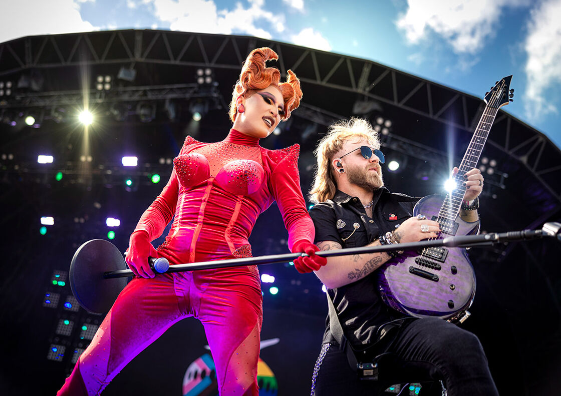 A drag queen in a bedazzled red suit holds a mic stand next to a guitarist 