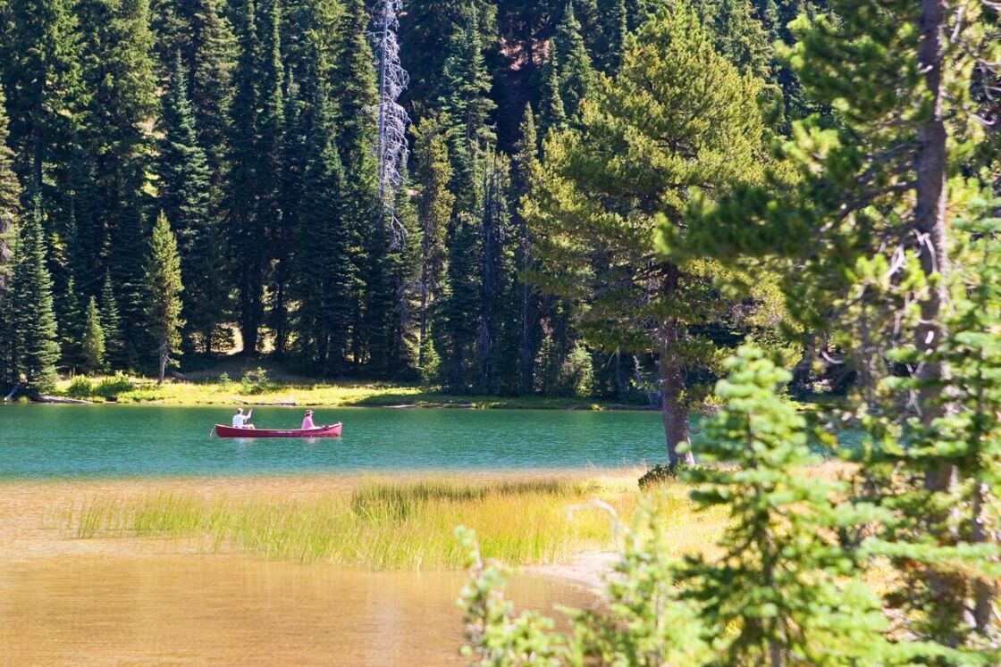 A canoe holding two people sits in the middle of a gorgeous lake, surrounded by evergreen trees