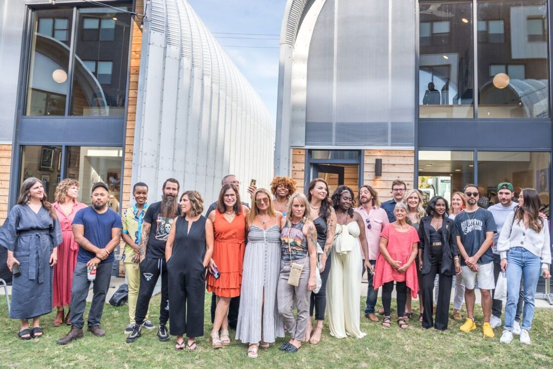 A large and diverse group of adults stands in front of large windowed hangars. Photo via City Goods