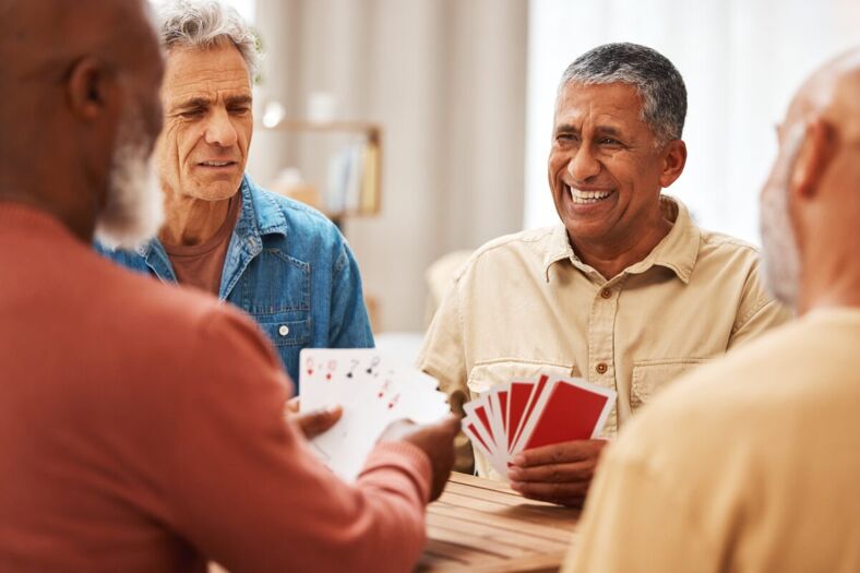 A group of four elderly men sit around a table playing cards. They are engaged in the game, with one man smiling broadly and the others focusing on their hands.