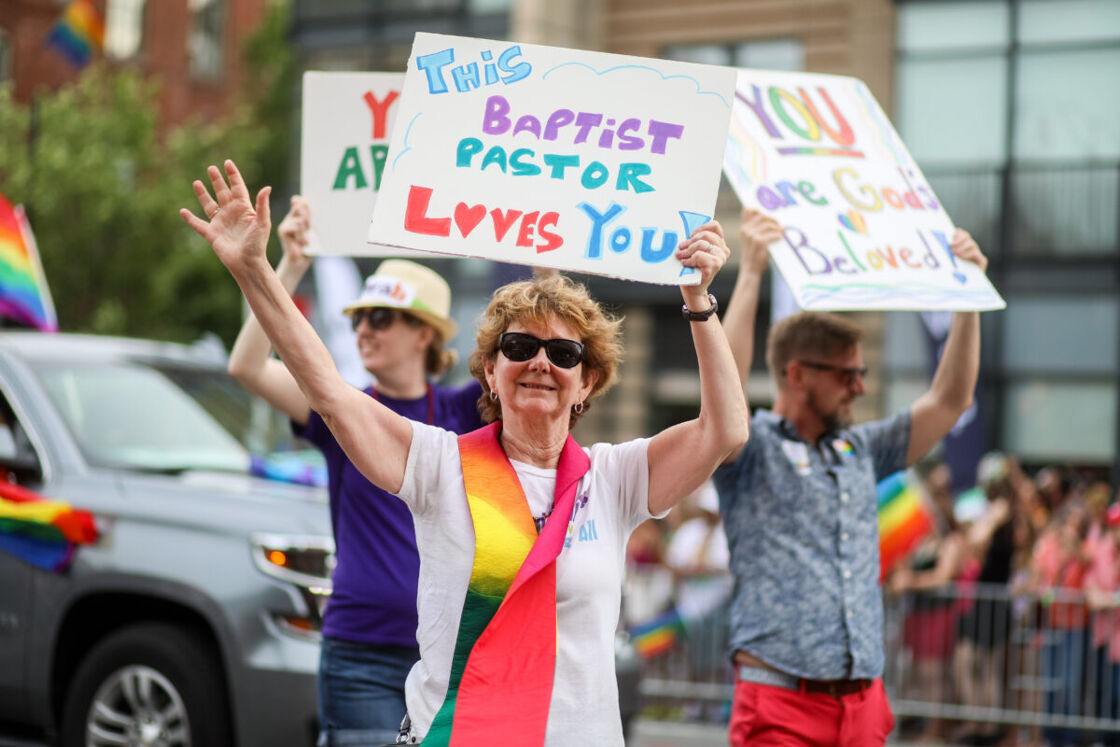 A female presenting person wearing a rainbow sash at a Washington D.C. Pride parade holding a sign that says 