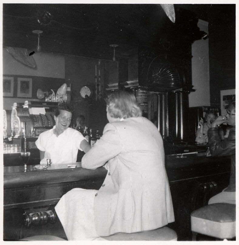 Grace Miller with white shirt behind the bar serving a woman with coat a drink. 