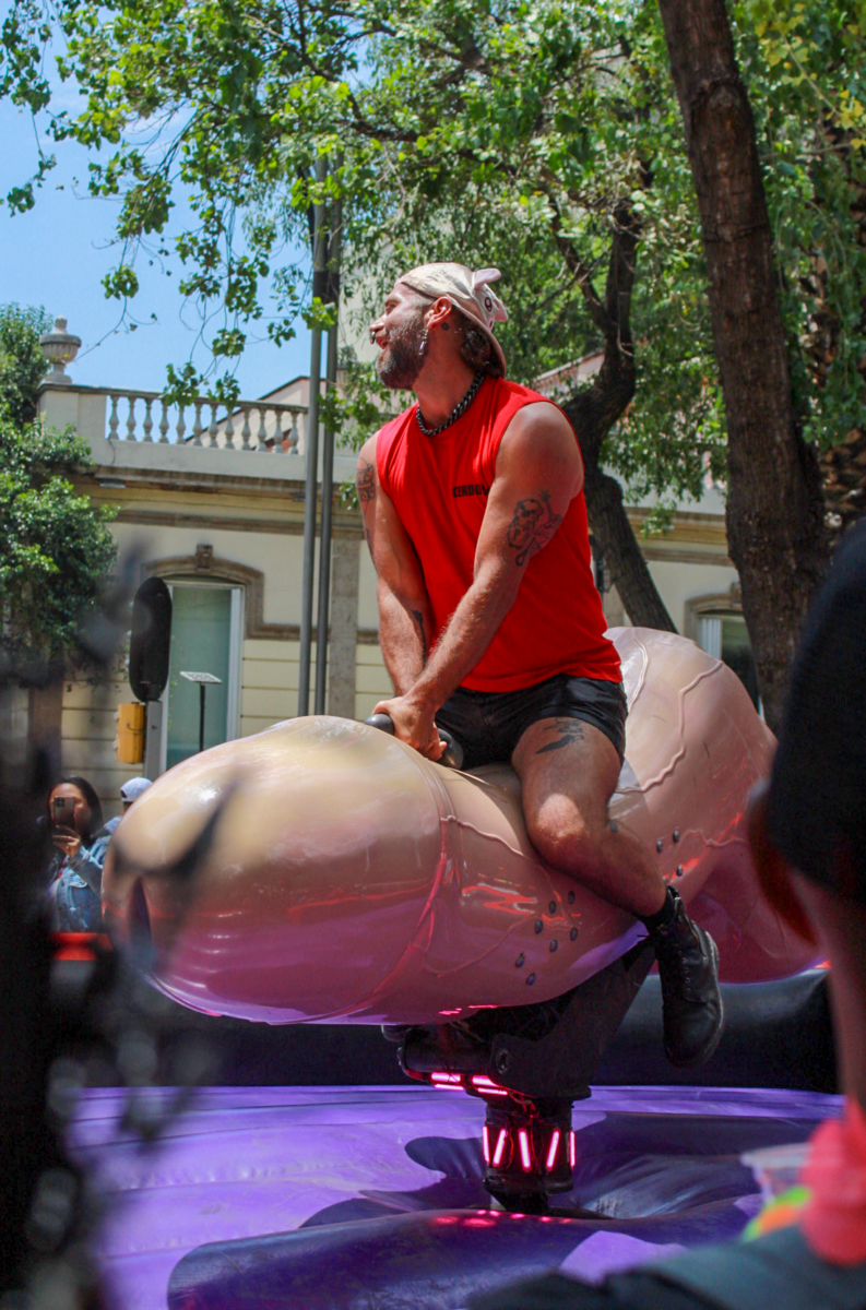 A man wearing a red shirt and black shorts rides a mechanical phallus ride