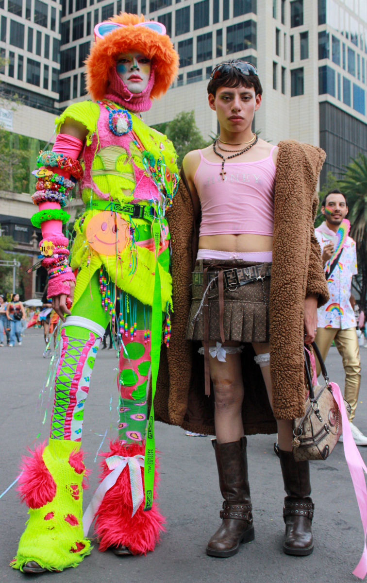 A person in a colorful yellow and green costume and wearing an orange wig stands next to a young nonbinary person wearing a brown coat and leather skirt.